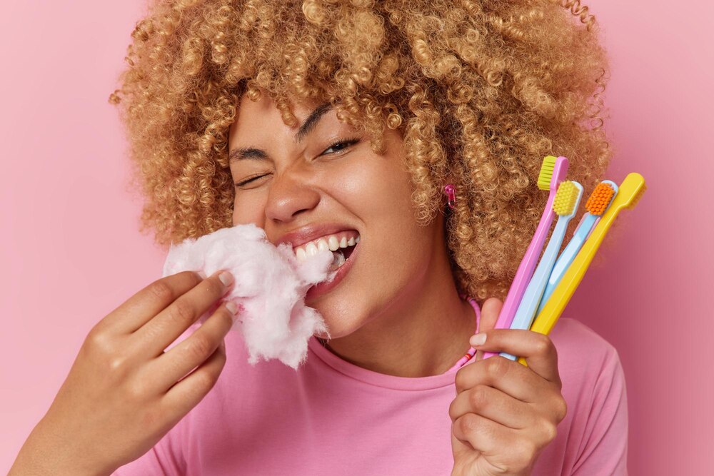 Horizontal shot of curly haired young woman bites appetizing candy floss harmful for teeth holds many toothbrushes in hand poses against pink background Sweet unhealthy food and sugar addiction