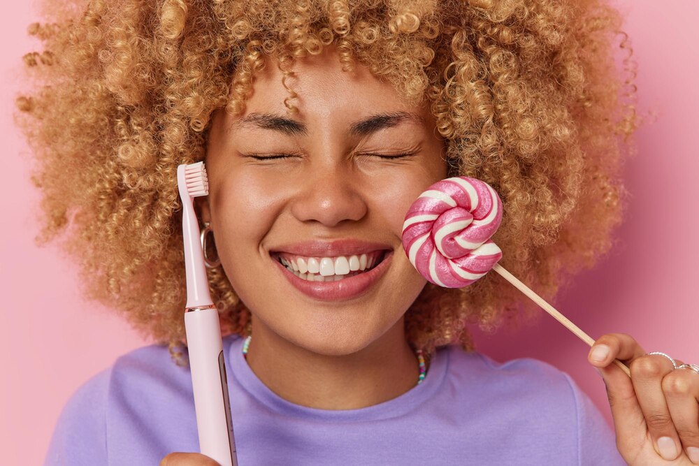 Headshot of cheerful curly haired woman keeps eyes closed smiles broadly has white even teeth holds caramel candy and electric toothbrush poses against pink background Harmful food concept
