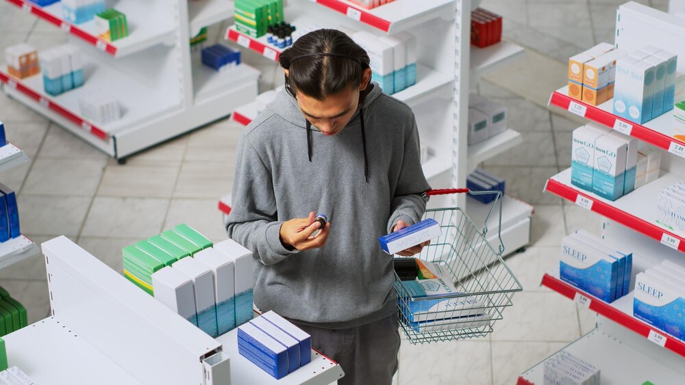 Young man looking at boxes of drugs in pharmacy shop, reading package to buy prescription medicine. Checking medicaments and supplements before taking treatment from shelves. Handheld shot.