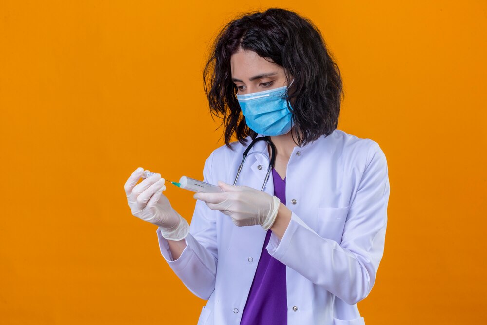 woman doctor wearing white coat with stethoscope in medical protective mask standing with syringe in hands looking at it with serious face on isolated orange